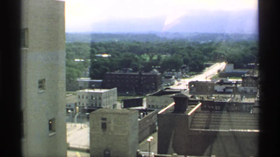 1970 Minnesota town view from above, with an adjacent wall, commercial rooftops, a sunlit street, trees, and distant low buildings.