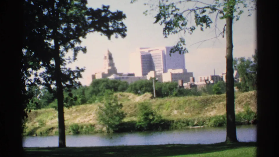 1970 8mm Minnesota park view with trees and grass in the foreground, water in the middle distance, and a city skyline beyond.