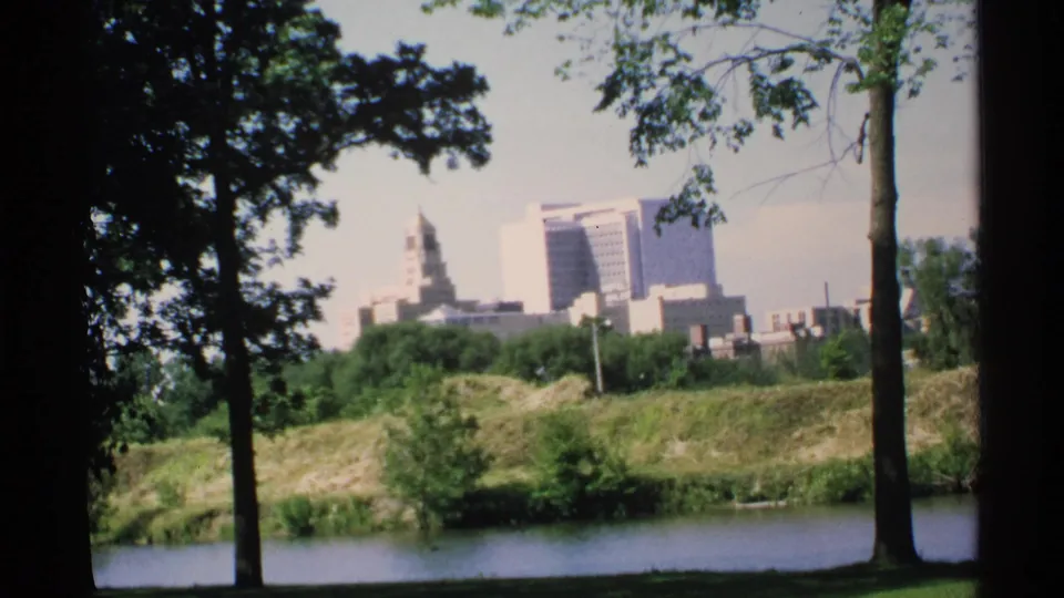 1970 8mm Minnesota frame looking from a shaded park across water and grass toward a distant skyline with a prominent towered structure.