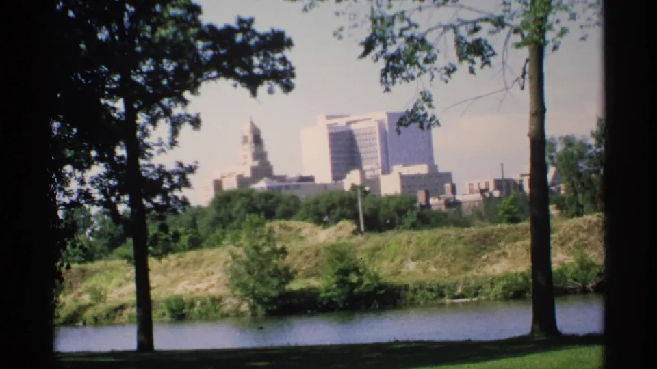 1970 8mm Minnesota home-movie view of a skyline across a pond, with tree trunks, shaded lawn, and a grassy embankment in front.