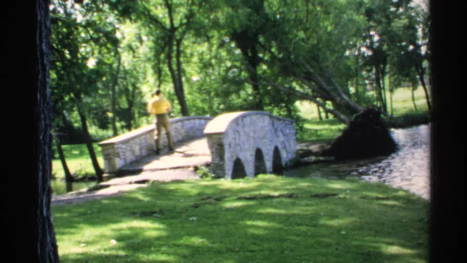 The bridge and stream remain in view as the man stands in bright sun within a wooded Minnesota park setting.