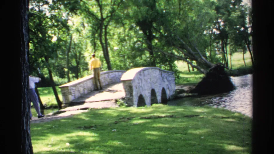 A second man enters at the left edge while the man in yellow remains on the stone bridge over the stream.