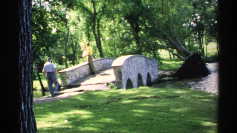 Two adult men stand near the small stone bridge in 1970 Minnesota home-movie footage of a leafy streamside park.