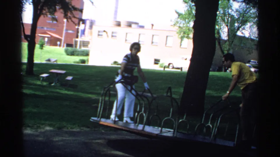Two adults use a metal seesaw in a shaded Minnesota park, with brick buildings and trees behind them.