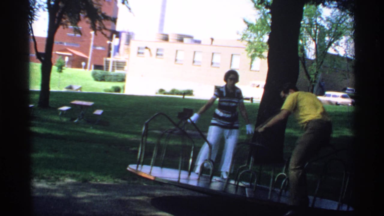 In 1970 Minnesota home-movie footage, two adults stand near a metal merry-go-round with trees, grass, and a brick building behind them.
