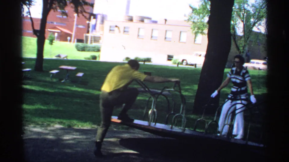 The man in a yellow shirt moves onto the metal merry-go-round while the woman in striped clothing watches from the side.