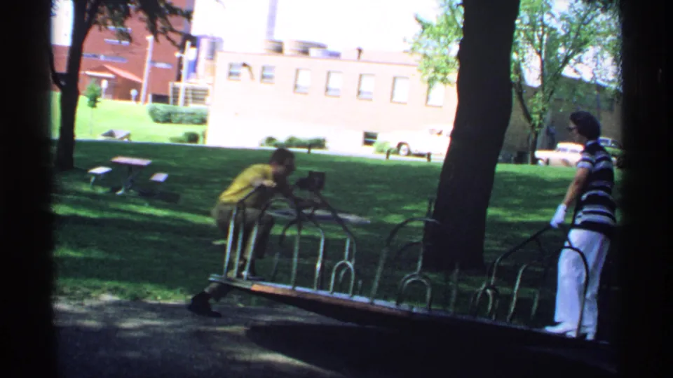 In the shaded park, the man leans onto the merry-go-round rails as the woman stands opposite him near the moving platform.