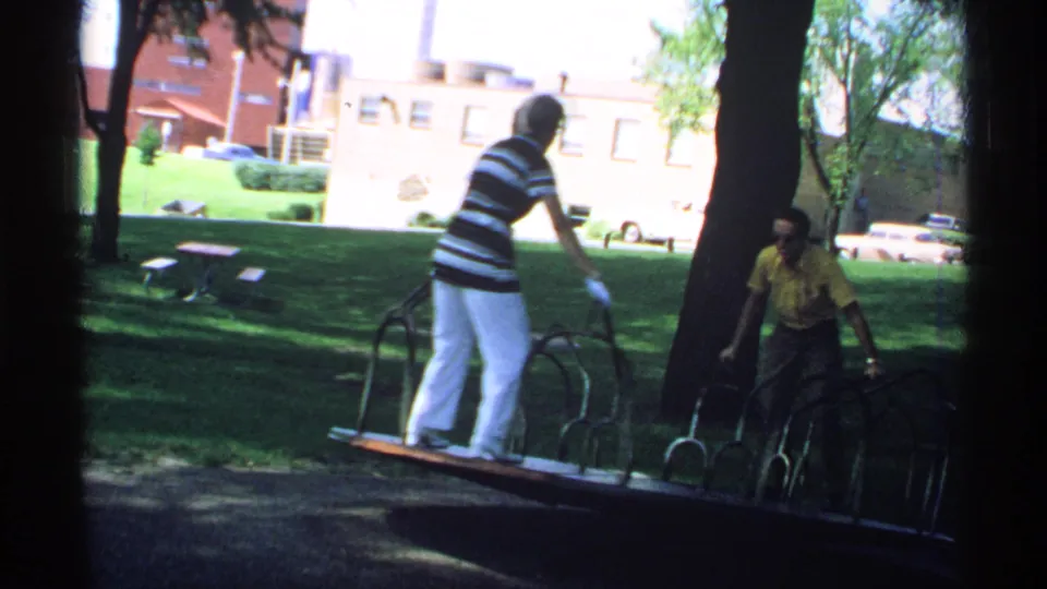 The woman in white pants stands on the merry-go-round platform as the man in a yellow shirt looks on in the park setting.