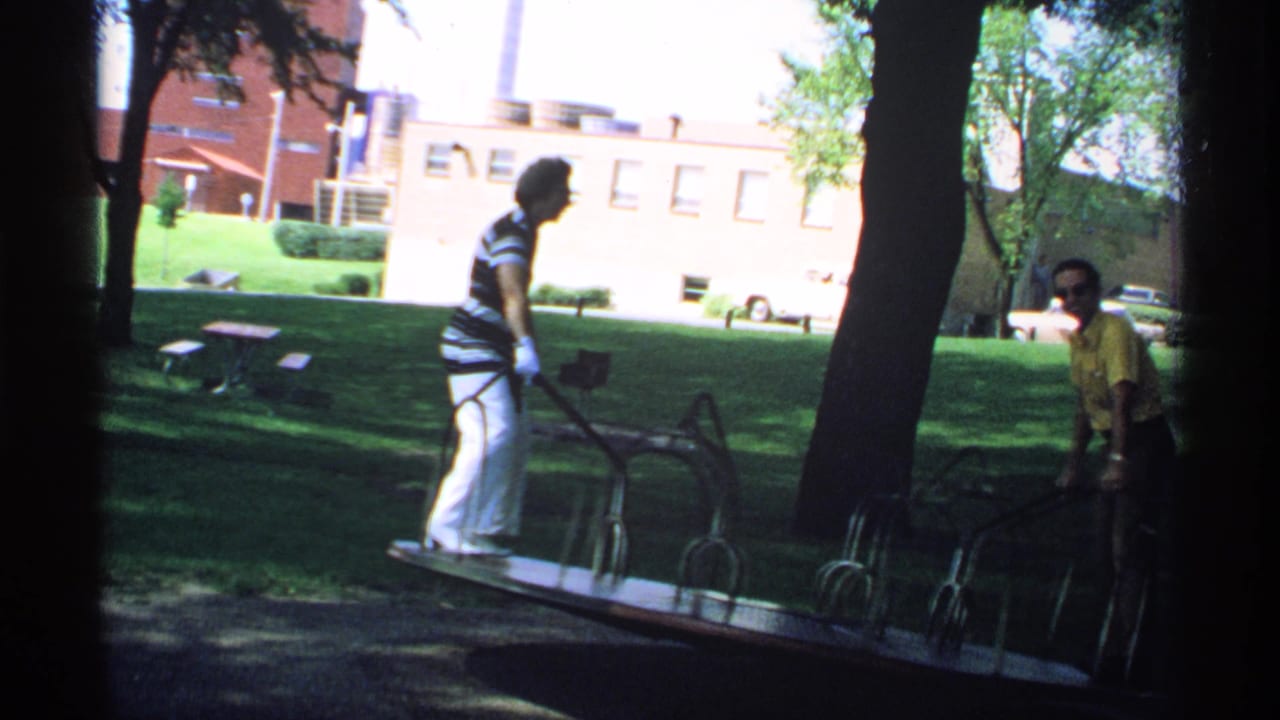 Woman in striped shirt stands on a park merry-go-round while a man in yellow sits or crouches nearby.