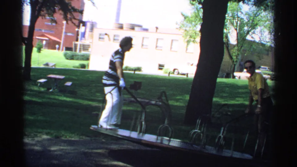 1970 Minnesota 8mm frame showing two adults at a shaded park merry-go-round with grass, trees, buildings, and parked cars beyond.