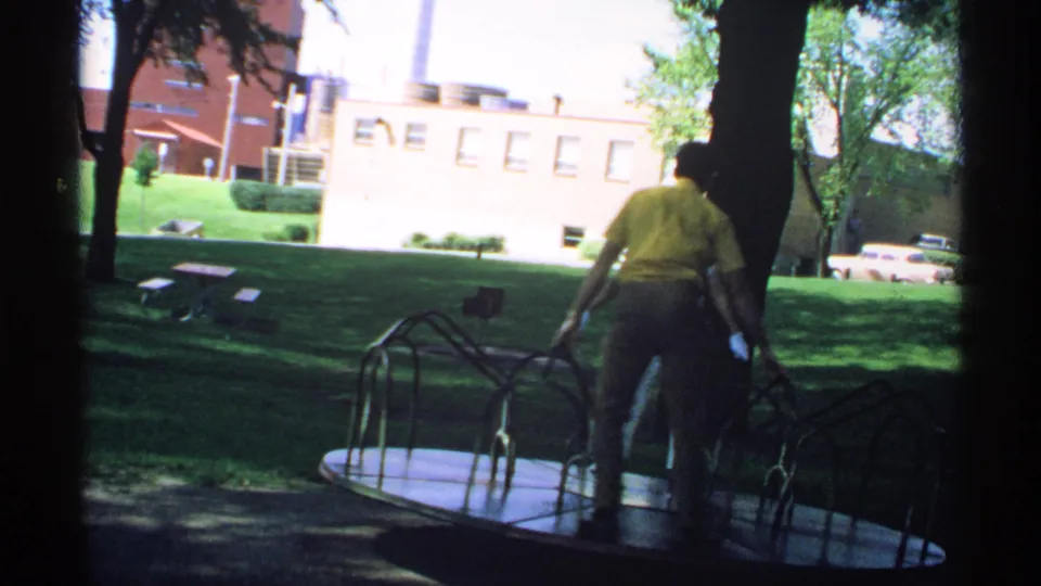 1970 Minnesota home-movie frame with a man in a yellow shirt near a metal merry-go-round, trees, lawn, buildings, and cars in view.