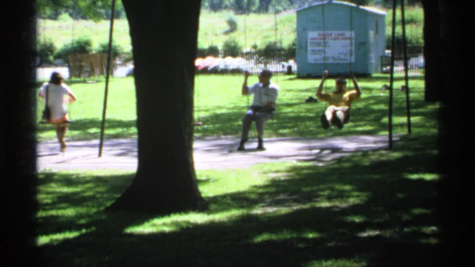 1970 Minnesota 8mm frame showing adults using playground swings, with large trees, grass, and a Silver Lake sign on a small building behind them.