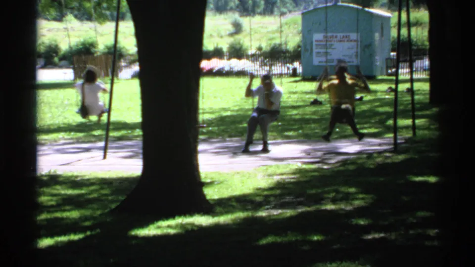 1970 Minnesota home-movie frame of adults on swings in a shaded park, with a small building marked Silver Lake in the background.