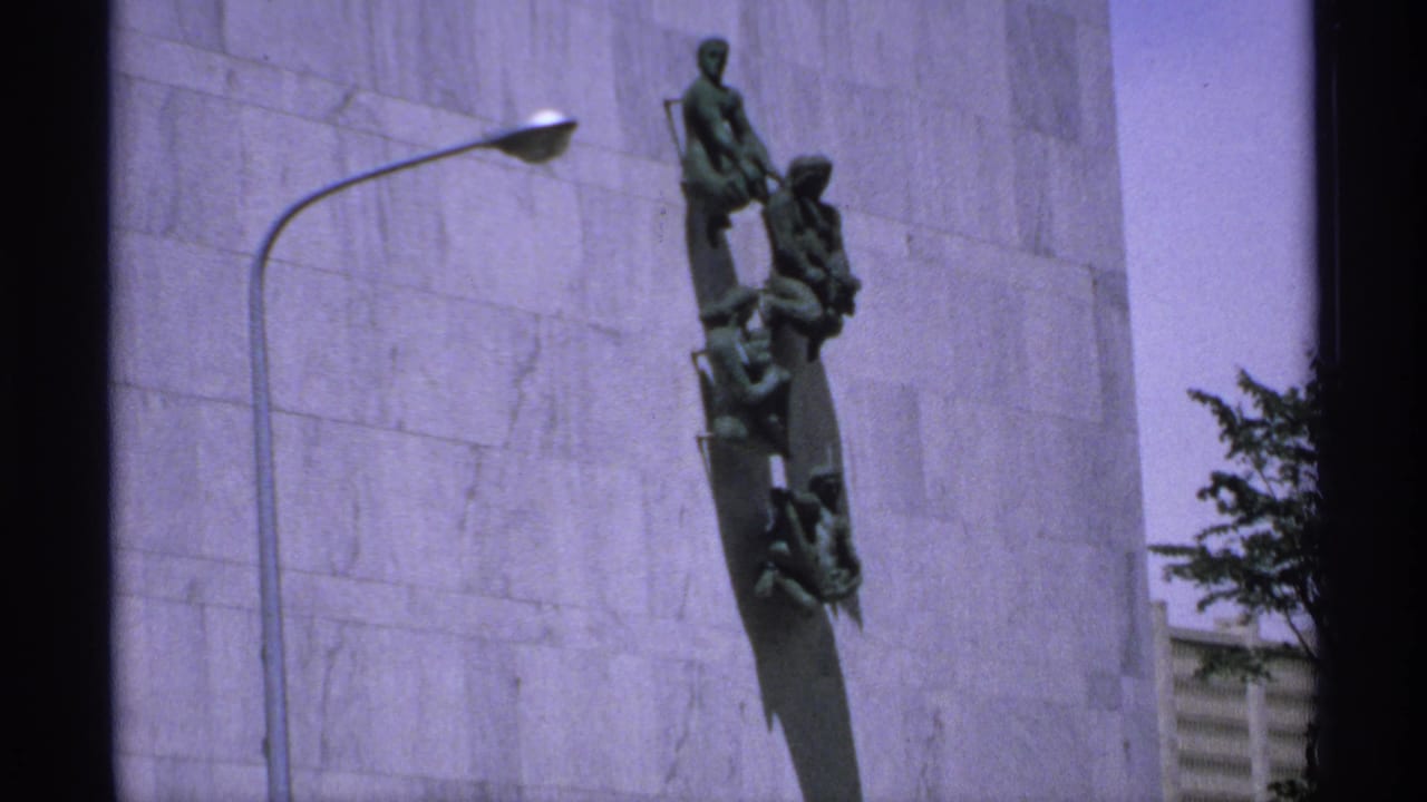 1970 Minnesota 8mm frame showing a green bronze vertical monument on a light stone facade, with a curved streetlamp at left.