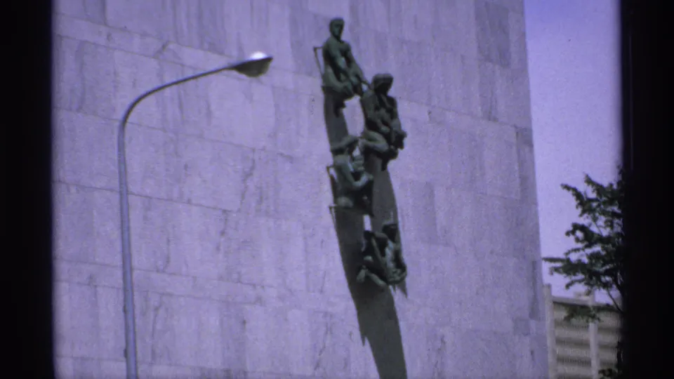 1970 Minnesota 8mm frame showing a green bronze vertical monument on a light stone facade, with a curved streetlamp at left.