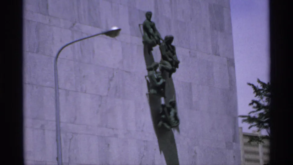 1970 Minnesota exterior frame with the bronze wall-mounted monument centered against pale stone and tree foliage near the right edge.