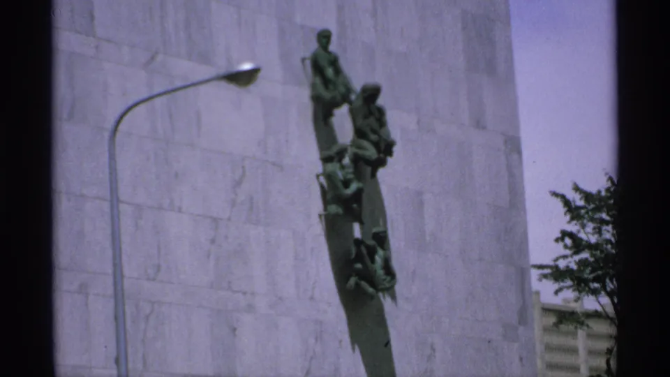 1970 Minnesota home-movie frame showing a handheld view of the green bronze monument mounted vertically on a stone building exterior.