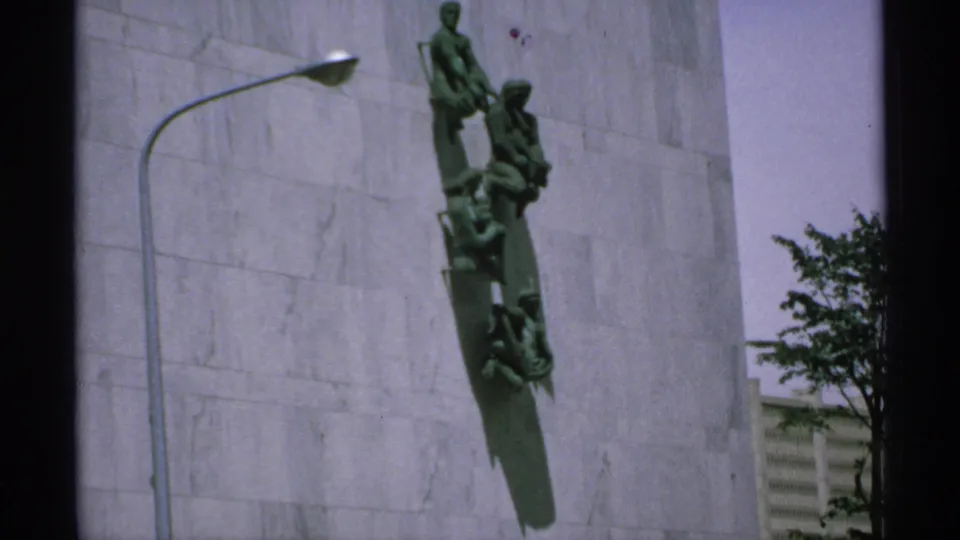 1970 Minnesota 8mm frame with the bronze mounted monument, long wall shadow, curved streetlamp, and dark tree foliage at right.