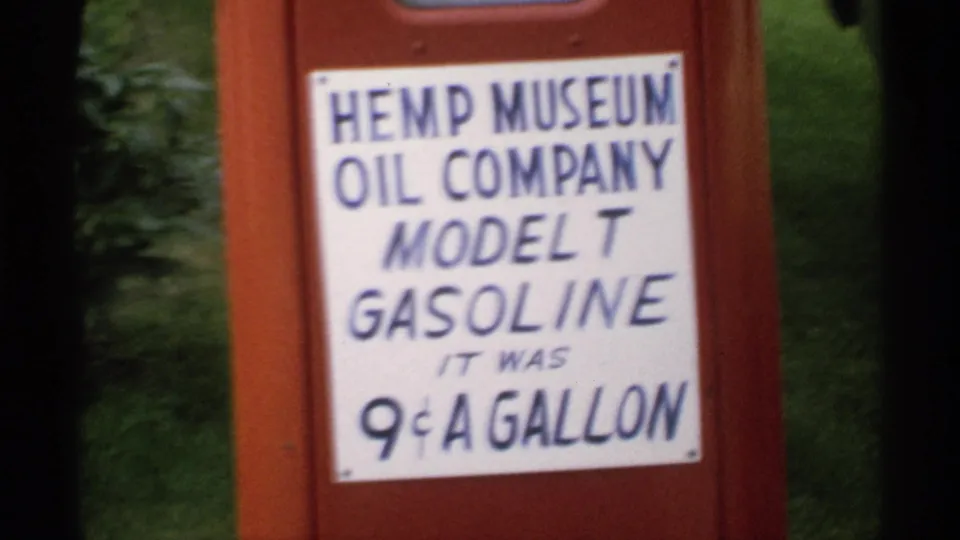 1970 Minnesota 8mm frame of a red pump display with a white Hemp Museum Oil Company Model T Gasoline sign against green foliage.