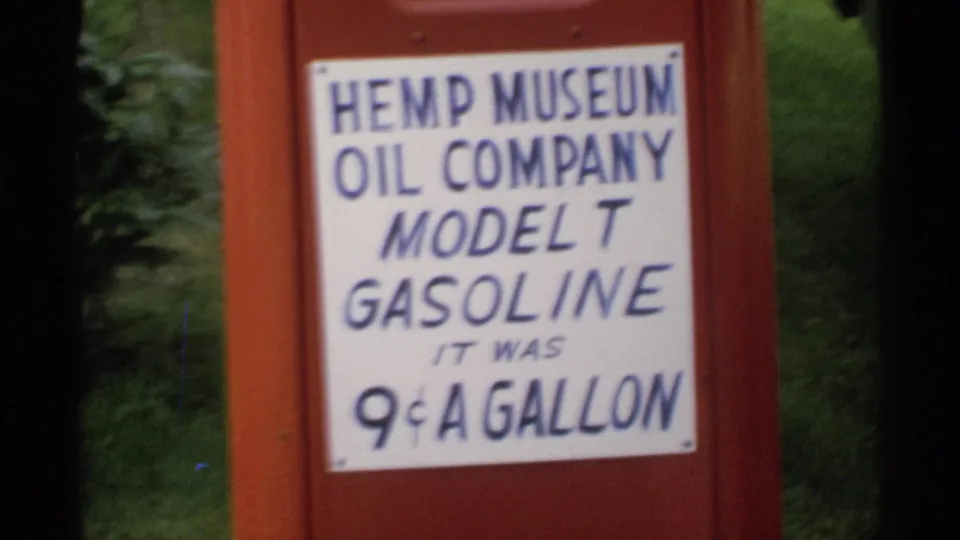 Soft 8mm close-up of the Hemp Museum Oil Company Model T Gasoline sign, with foliage visible beside the red housing.