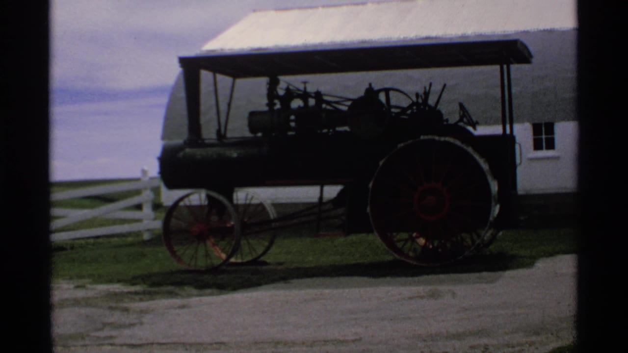 Canopy-topped steam tractor beside a white barn and fence.