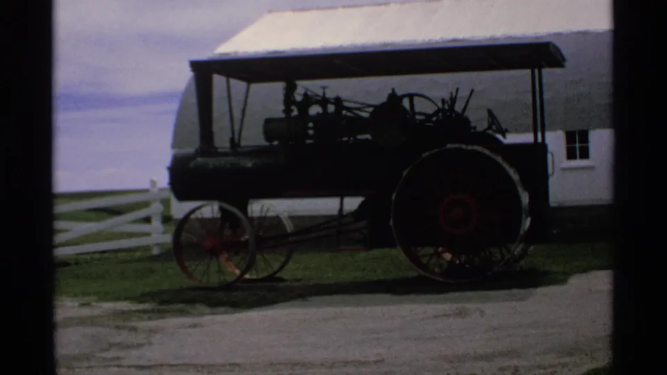 Minnesota 1970 8mm frame showing a vintage steam tractor with large red-spoked wheels near a white barn and rail fence.