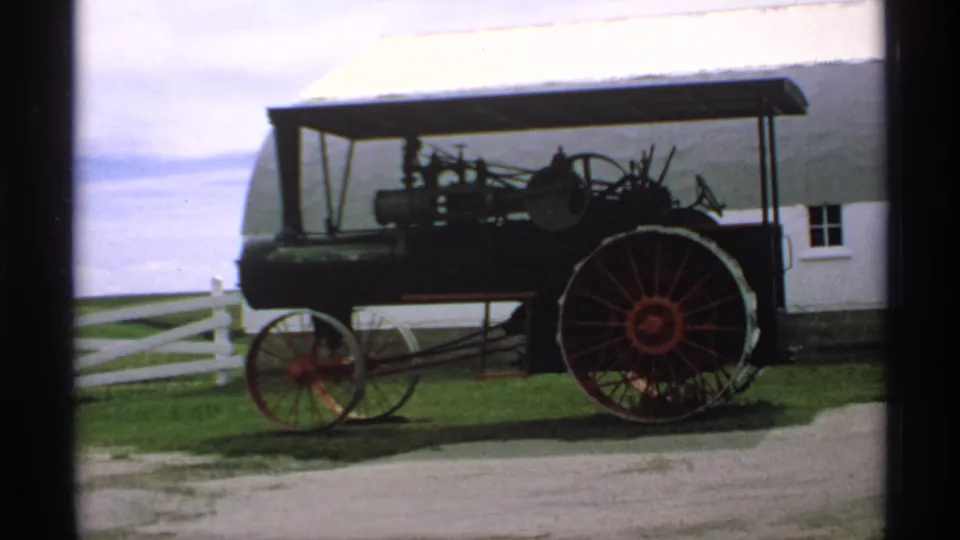 A vintage steam tractor with a canopy and red wheels sits on grass near a white barn, consistent with the Minnesota 1970 clip metadata.