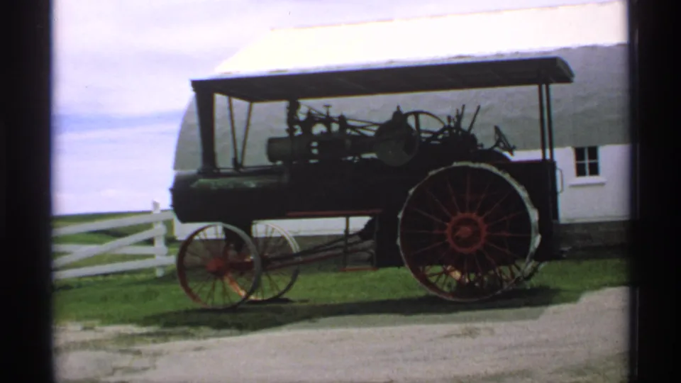 1970s 8mm view of an antique steam tractor display, with red-spoked wheels, white barn siding, grass, and a rail fence visible.