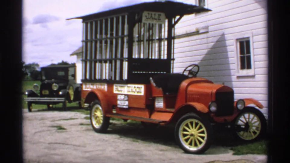 Minnesota 1970 frame showing a red early delivery-style truck with cage-like cargo area and yellow wheels parked beside a white building.