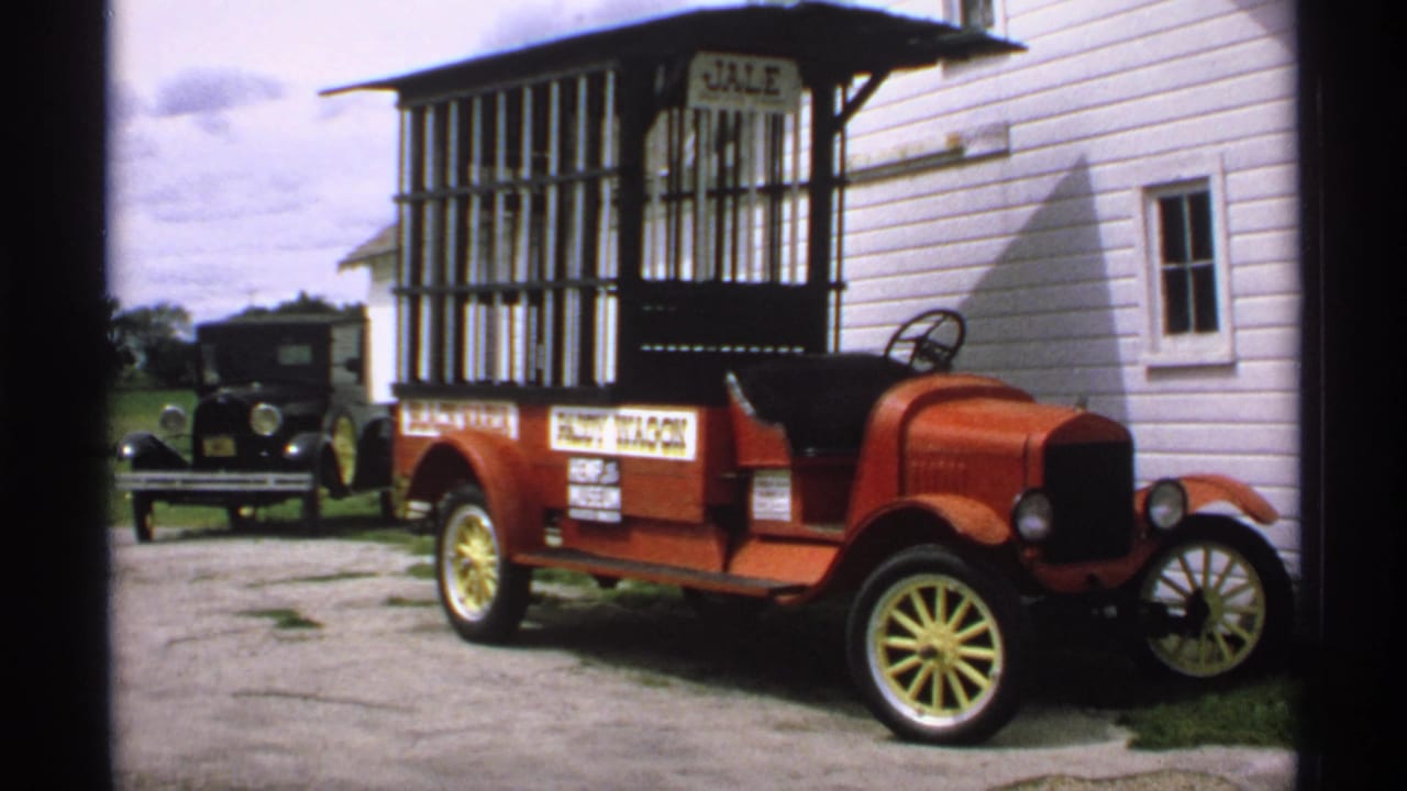 Red open-top vintage-style car parked beside a white clapboard building.