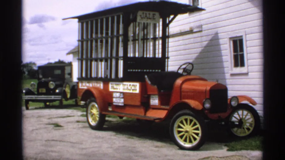 The red JALE-labeled open-top car remains parked in daylight, with a black older automobile visible behind it near the building.