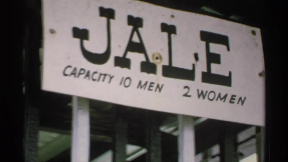 1970 Minnesota home-movie frame showing a weathered white JALE sign with capacity wording for men and women.