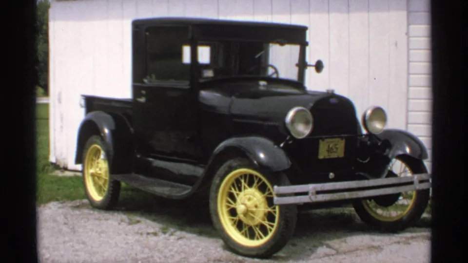 1970 Minnesota home-movie frame showing a black vintage pickup, identified in metadata as a Ford Model T, beside a white building.
