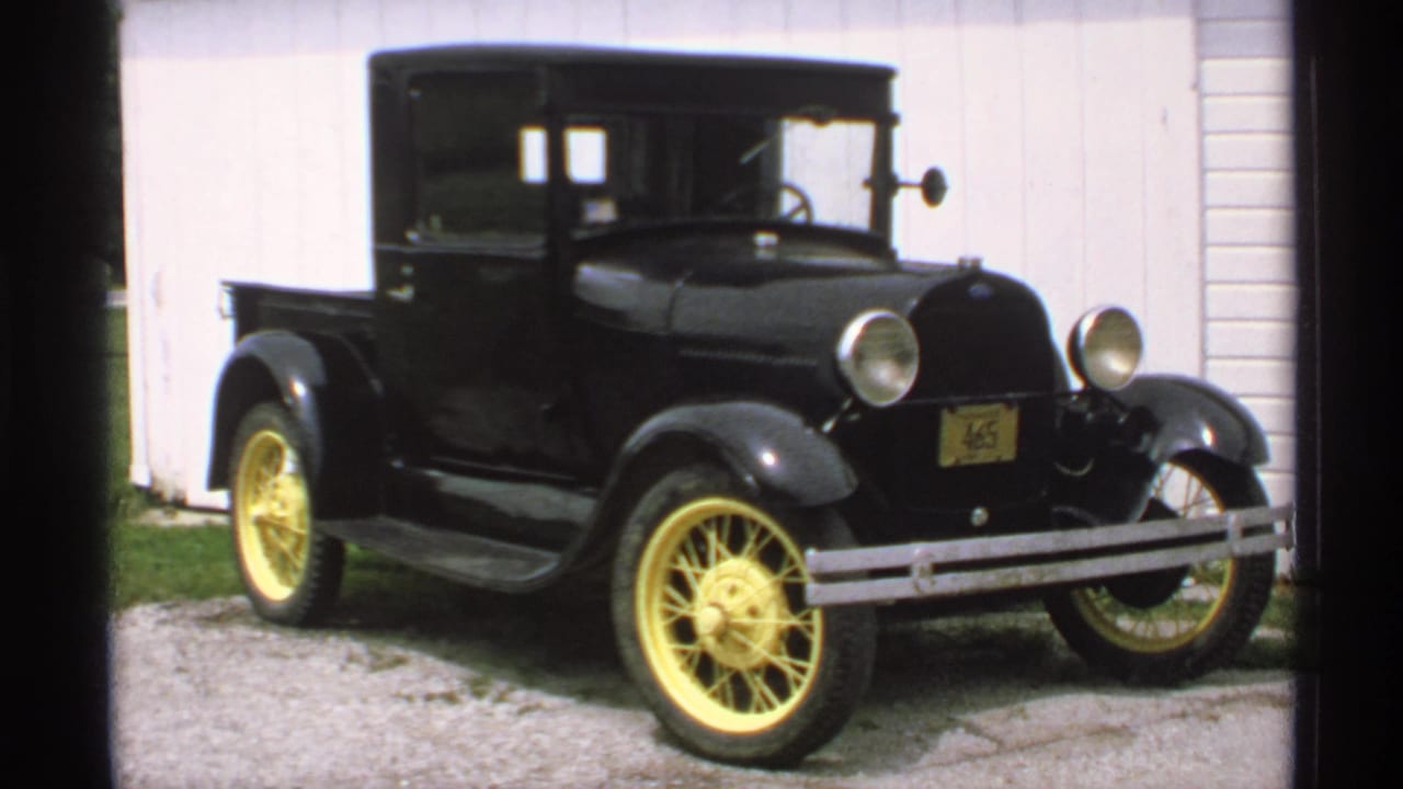 1970 Minnesota 8mm frame of a black vintage Ford pickup with yellow wheels on a gravel drive beside a white building.