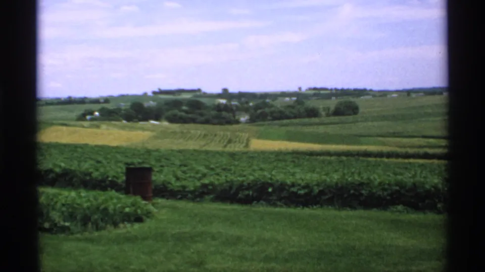 1970 Minnesota 8mm frame of a quiet farm landscape with foreground lawn, cultivated fields, distant trees, and visible film-frame edges.