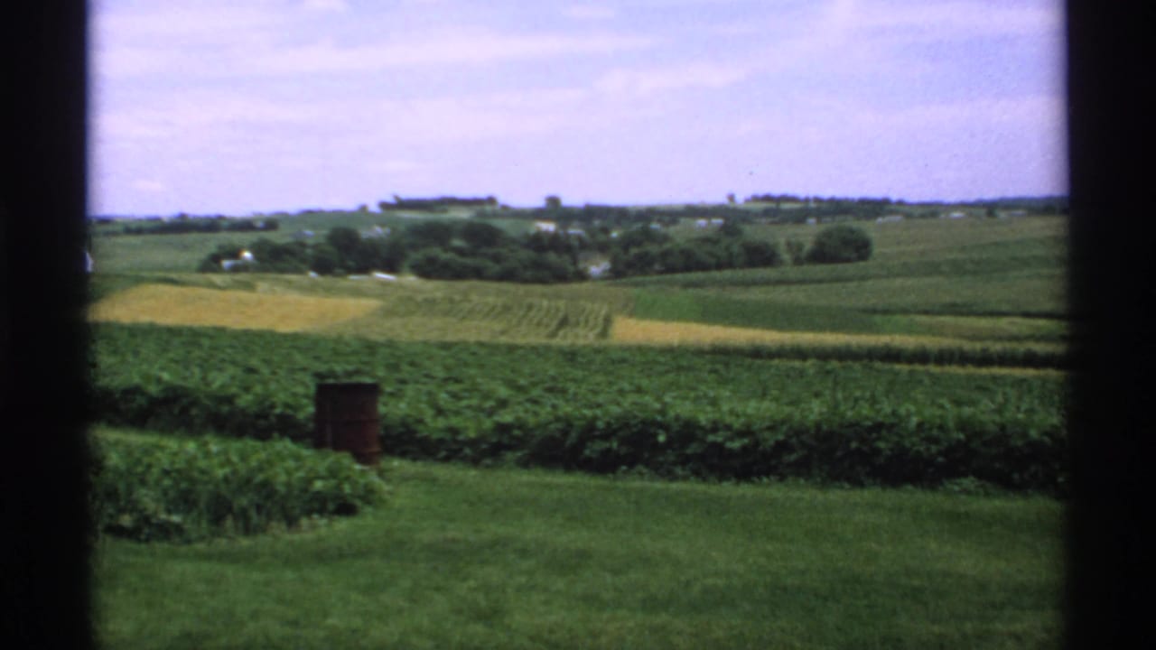 Rolling green farmland seen through dark window borders with a small red barrel in the foreground.