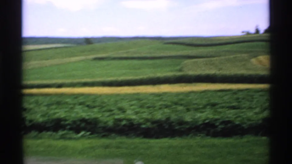 Soft 8mm frame of Minnesota crop fields, rolling green hills, and a yellow field band viewed through a dark opening.
