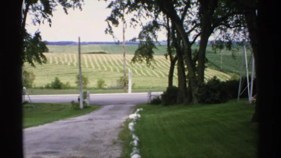 1970 Minnesota home-movie frame of a rural driveway, roadside fence posts, shade trees, and striped crop fields in the distance.