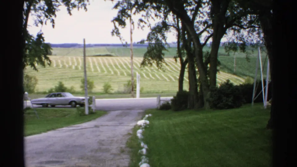 A silver sedan drives past the end of a rural Minnesota driveway, with 1970s home-movie color and fields beyond the road.