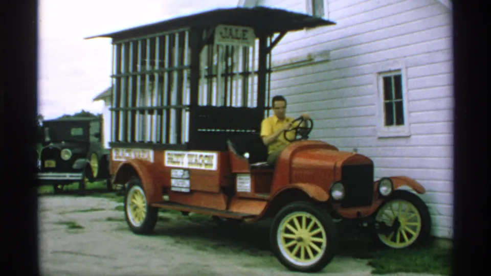 A man operates or poses in a red antique delivery truck with display signage beside a white clapboard building in 1970 Minnesota footage.