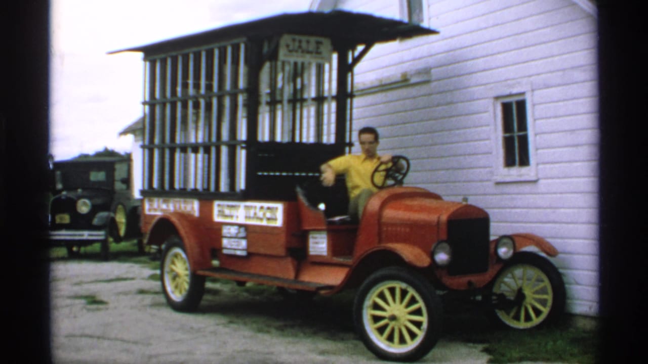 Man in a yellow shirt seated in a red vintage wagon car beside a white clapboard building.