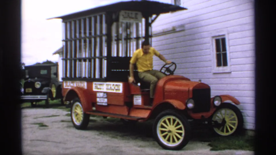 The man shifts out of the driver's seat of the red vintage wagon car, with signage and a black vehicle visible in the background.