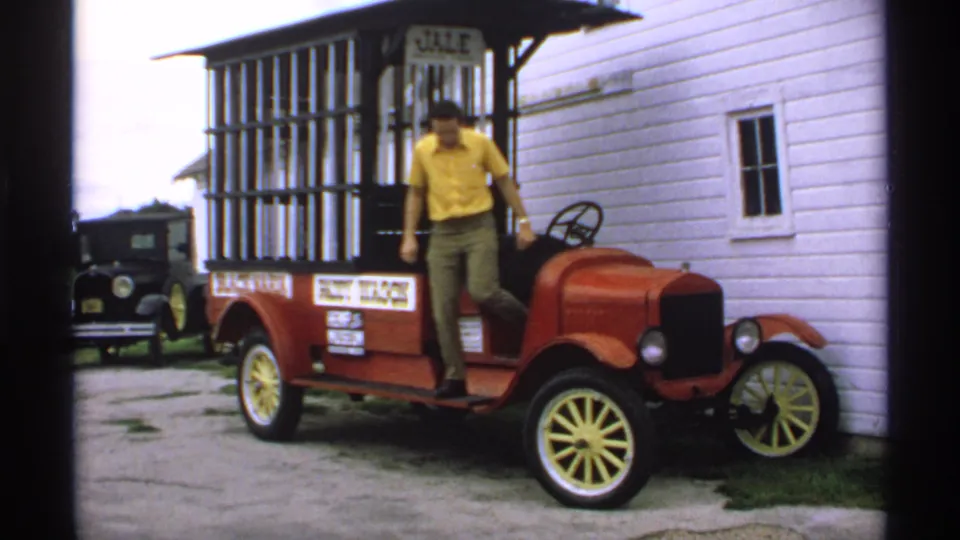 The man steps from the red vintage fruit wagon car onto the gravel area beside the white clapboard building in Minnesota home-movie footage.