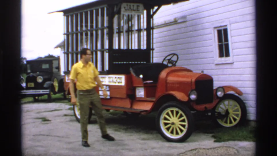 After leaving the driver's seat, the man stands beside the red vintage wagon car, with the white building and black background car still visible.