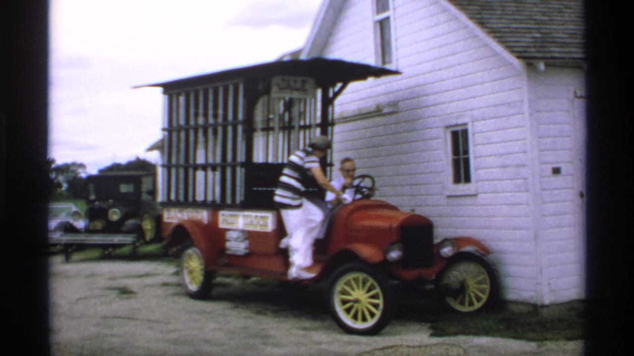 Woman climbing into a red antique paddy wagon beside a white clapboard building.