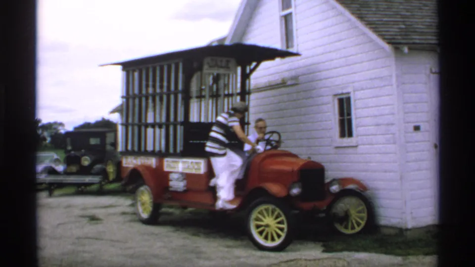 In 1970s Minnesota 8mm footage, a woman climbs into a red antique paddy wagon parked beside a white clapboard building with a JAIL sign.