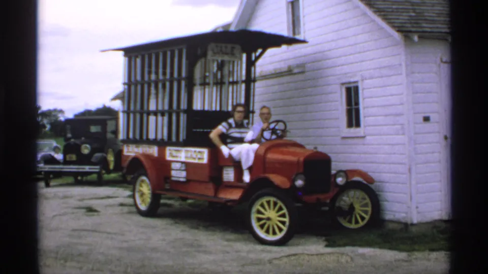 Two adults sit in the front of the red antique paddy wagon, with its barred rear compartment, yellow wheels, and white clapboard building visible.