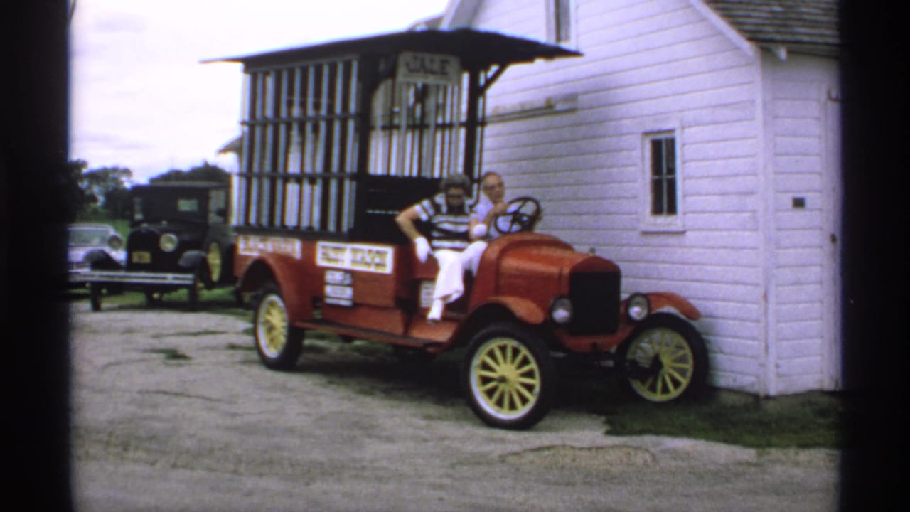 Two adults sit in a red antique-style sales car beside a white clapboard building.