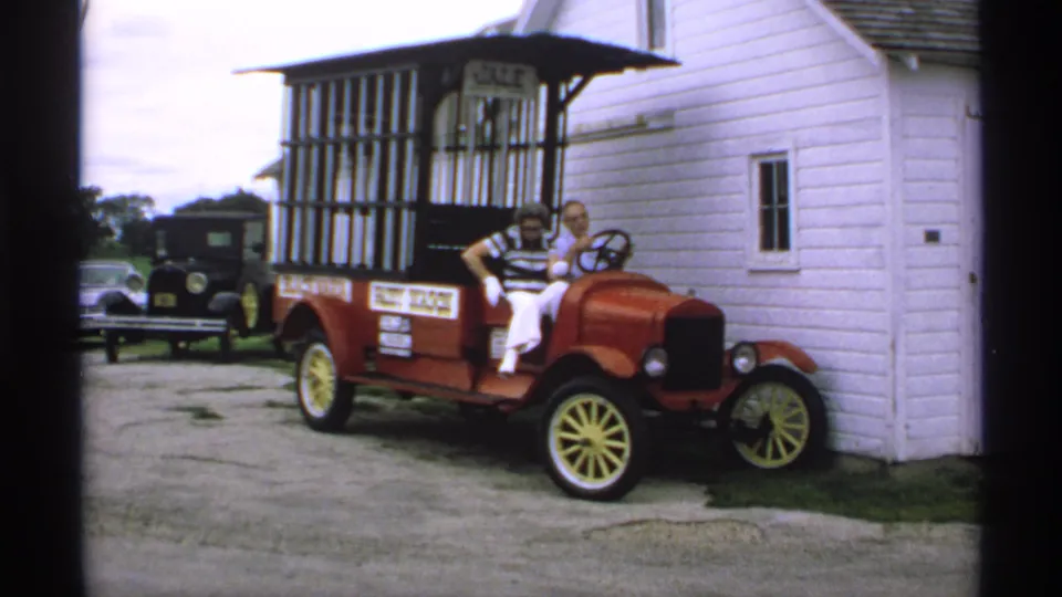 In 1970s Minnesota 8mm footage, a couple sits in a red Model T-style sales car with yellow wheels near a white clapboard building.