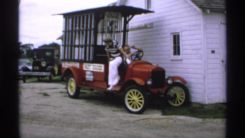 The woman in a striped top and white pants exits the red sales car while the seated man stays by the wheel in Minnesota footage from 1970.